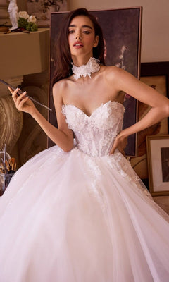 A woman poses indoors holding a paintbrush, wearing the Andrea and Leo WL041 long wedding dress with a strapless floral lace bodice, full A-line tulle skirt, and matching lace choker.