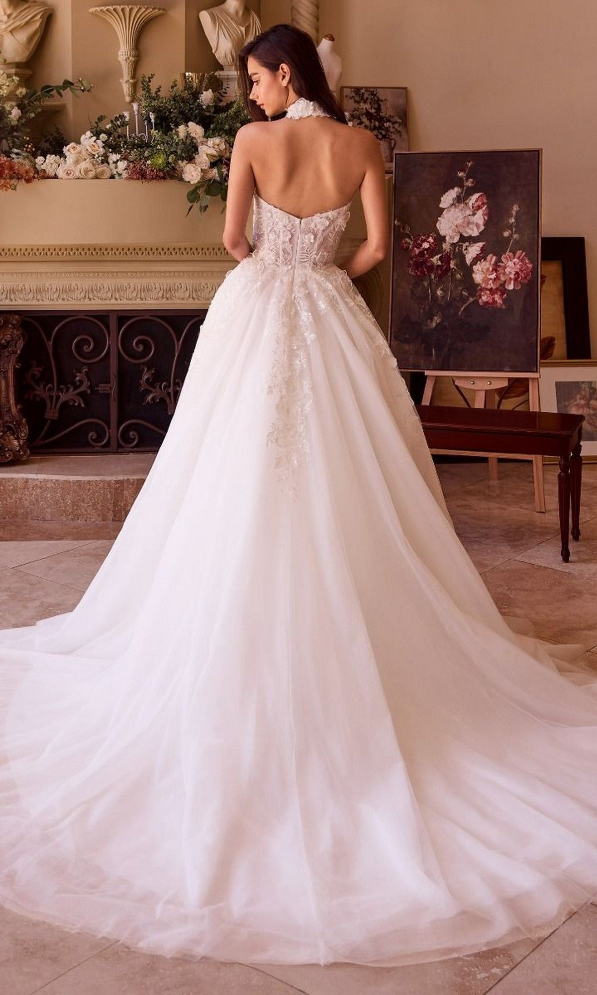 A woman stands indoors wearing the Long Wedding Dress: Andrea and Leo WL041, a strapless gown with a floral lace bodice and flowing train; floral arrangements and framed artwork are visible in the background.