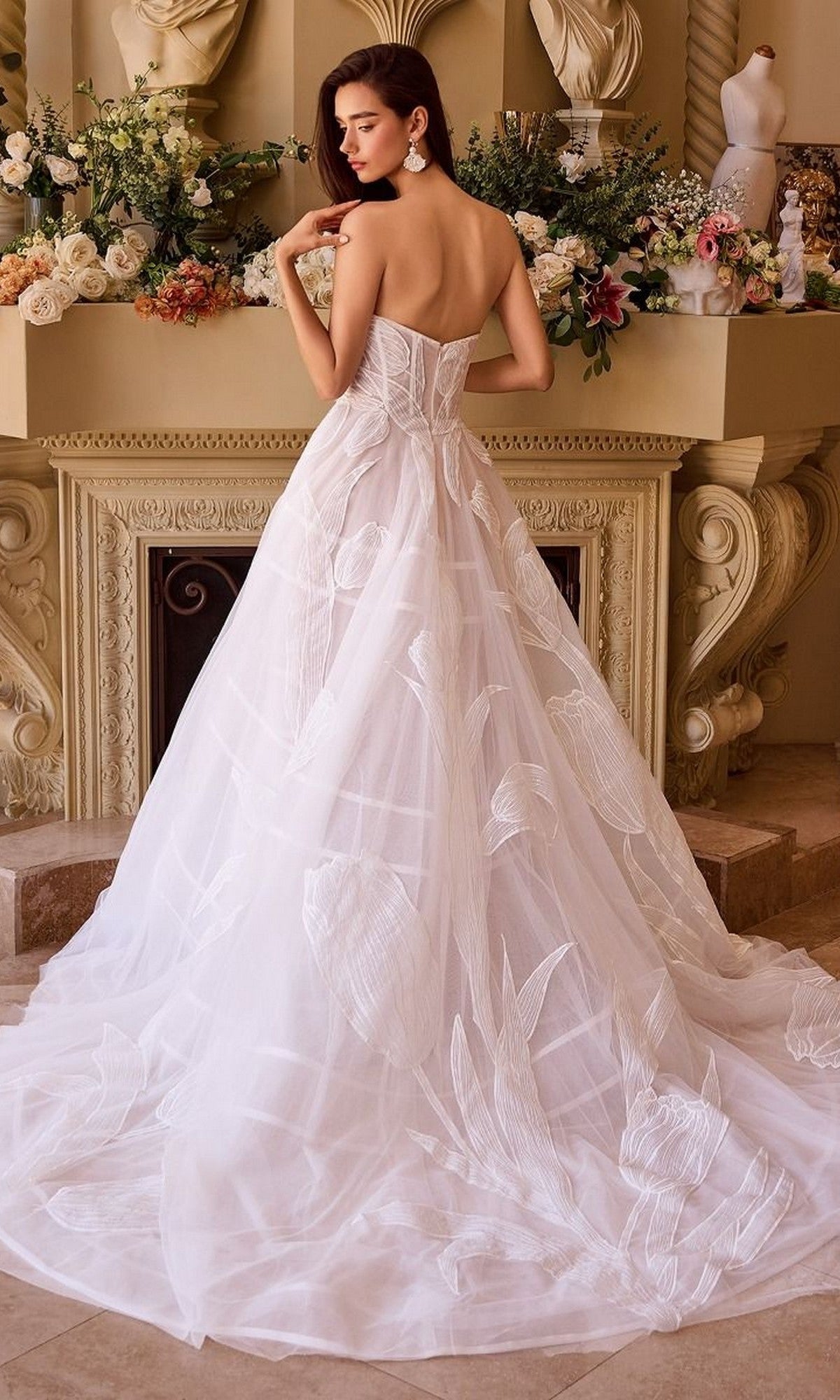 A woman wearing the Andrea and Leo WL043 long wedding dress with a strapless sweetheart neckline and tulle A-line skirt stands before a fireplace decorated with floral arrangements.