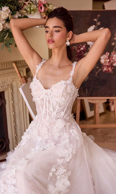 A woman in the Andrea and Leo WL045 long wedding dress with a detailed white floral design and lace corset bodice poses indoors, arms raised, surrounded by floral arrangements and a painting.
