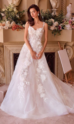 A woman in the Andrea and Leo WL045 long wedding dress with a lace corset bodice stands indoors before a flower-adorned mantle and an empty easel.
