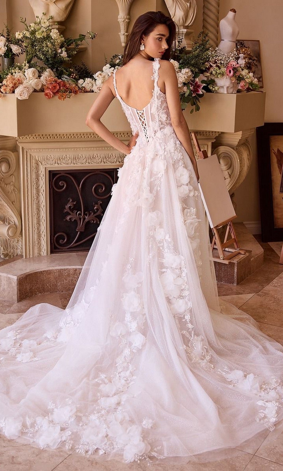 A woman in the Andrea and Leo WL045 long wedding dress with a lace corset bodice and flowing tulle A-line skirt stands indoors near a decorated mantel, looking over her shoulder.