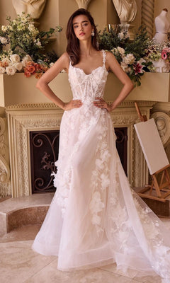 A woman stands before a fireplace wearing the Andrea and Leo WL045 long wedding dress, featuring a white floral lace corset bodice and flowing train. Elegant flower arrangements and decor complete the refined backdrop.