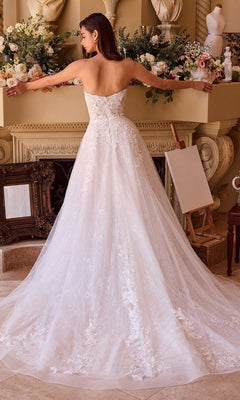 A woman wearing the Andrea and Leo WL052 long wedding dress, a white strapless gown with delicate floral lace applique, stands with arms outstretched in front of a flower-adorned fireplace mantel.