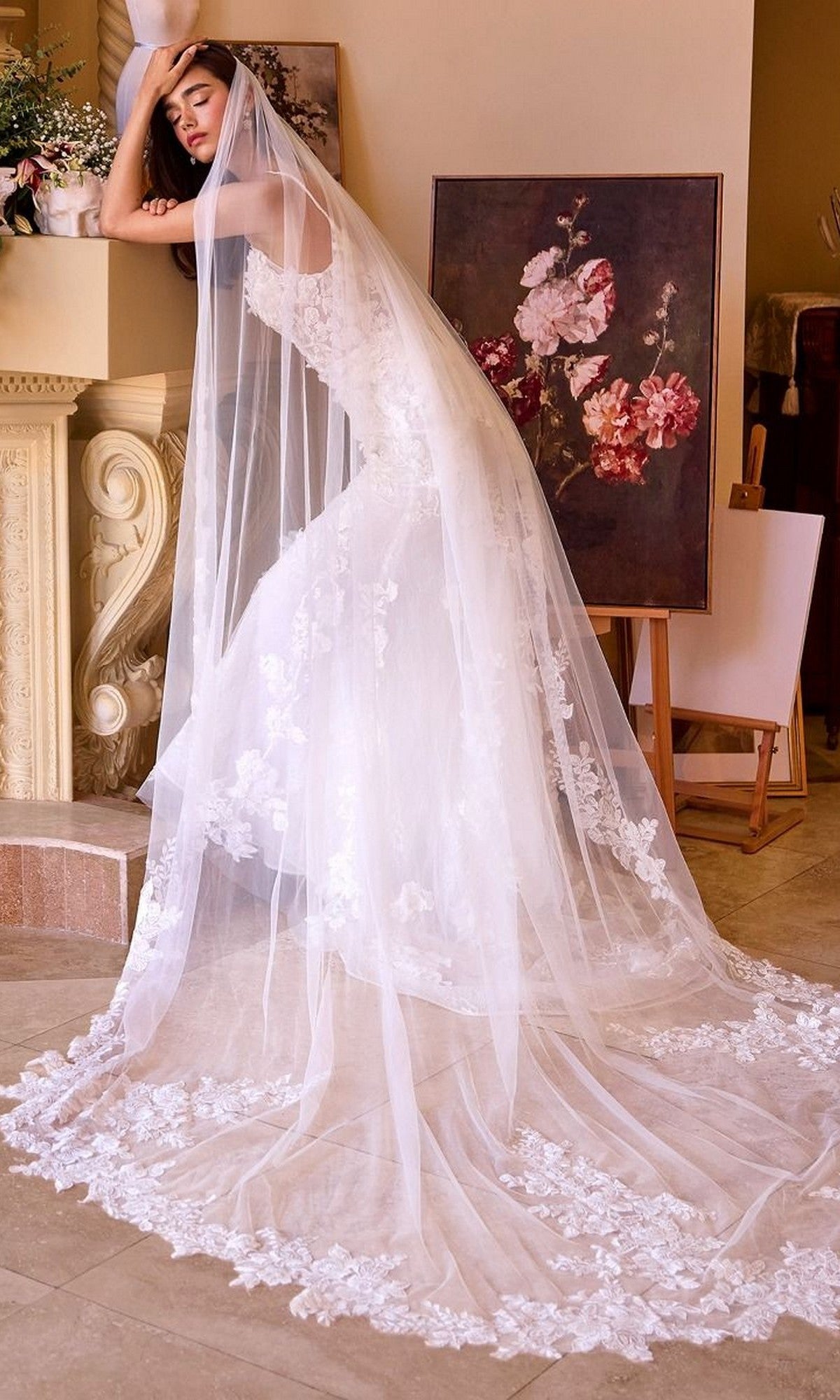 A woman in the Andrea and Leo WL053 long wedding dress, featuring sleeveless floral lace applique and a sheer veil, poses indoors beside a floral painting and ornate fireplace.