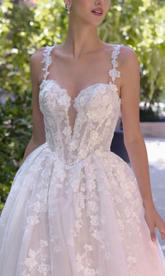 A woman stands outdoors amid greenery wearing the Andrea and Leo WL058 long wedding dress, featuring white floral lace, a deep neckline, spaghetti straps, and a full A-line skirt.