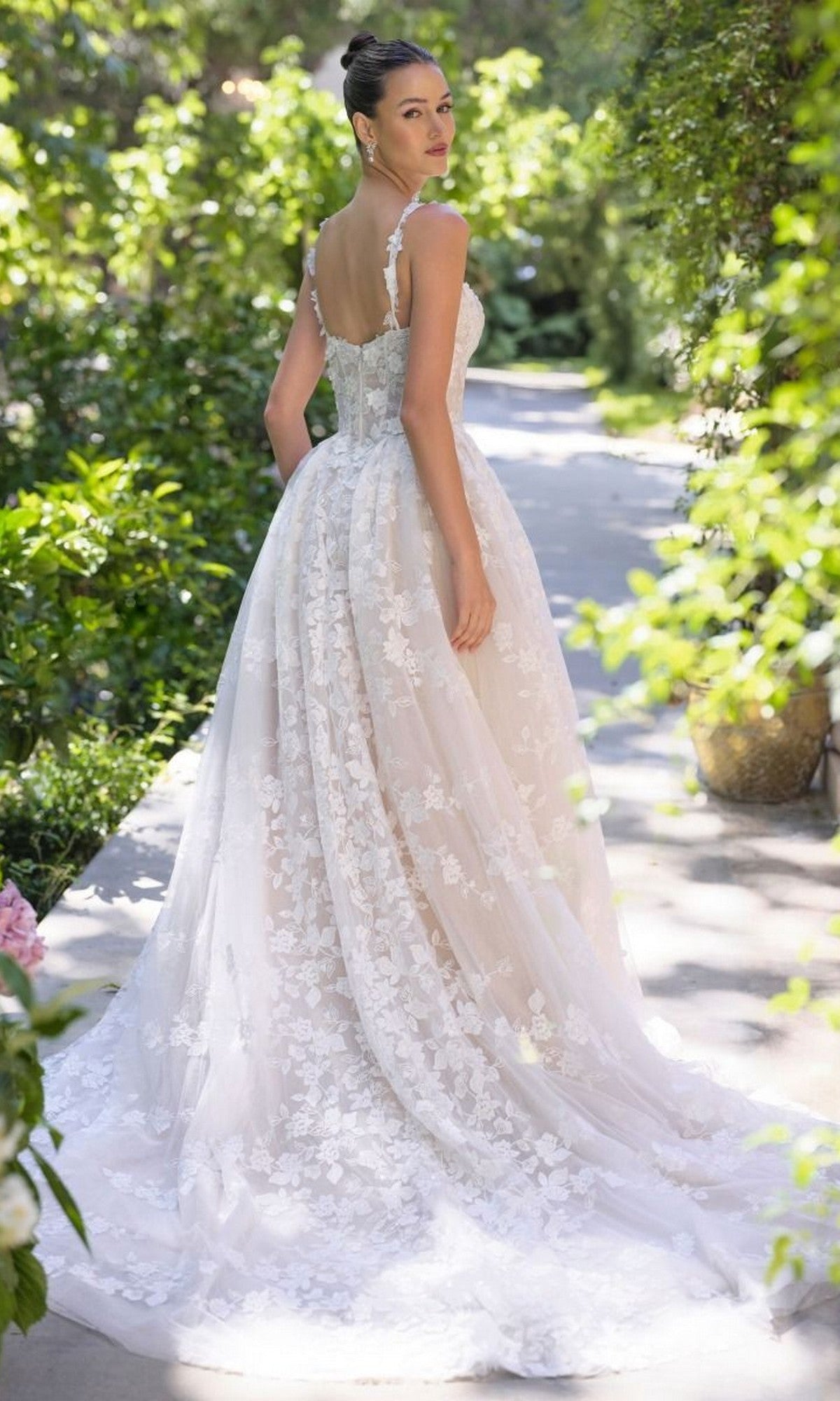 A woman stands in a garden wearing the Andrea and Leo WL058 long wedding dress, featuring a white floral lace A-line design with a full skirt and sheer lace train, as she looks back over her shoulder.