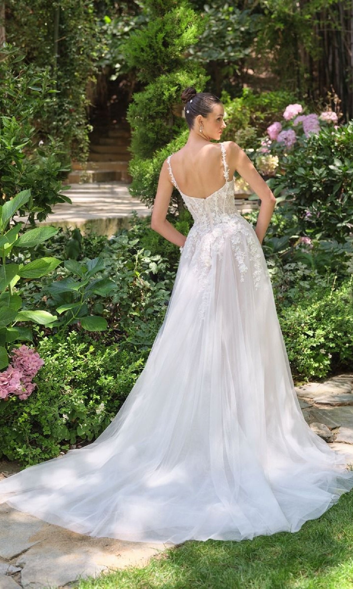 Woman showing the back of a white wedding dress she's wearing standing in a garden with greenery and flowers.