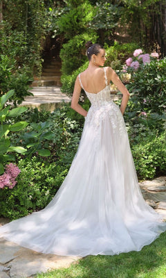 Woman showing the back of a white wedding dress she's wearing standing in a garden with greenery and flowers.