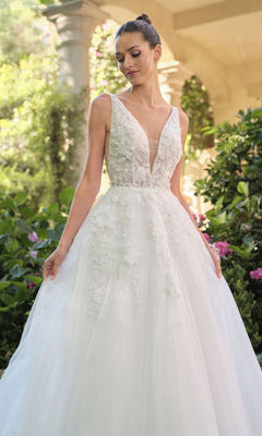 A woman stands outdoors among greenery and flowers wearing the Andrea and Leo WL064 long, white, sleeveless V-neck A-line wedding dress with floral embroidery and lace appliques.