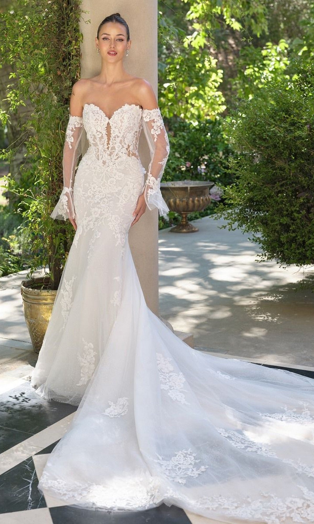 A woman stands outdoors on a sunlit patio wearing the Andrea and Leo WL065, a mermaid silhouette off-the-shoulder white lace wedding dress with detachable lace sleeves and a long train.