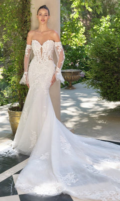 A woman stands outdoors on a sunlit patio wearing the Andrea and Leo WL065, a mermaid silhouette off-the-shoulder white lace wedding dress with detachable lace sleeves and a long train.
