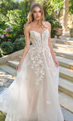 A woman stands outdoors on stone steps surrounded by lush greenery and vibrant flowers, wearing the Andrea and Leo WL070 long wedding dress with 3D floral lace appliques and a strapless neckline.