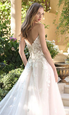 A woman with long, wavy hair stands outdoors wearing the Andrea and Leo WL070 Long Wedding Dress, which features 3D floral lace appliques and a low back. Greenery and flowers can be seen in the background.
