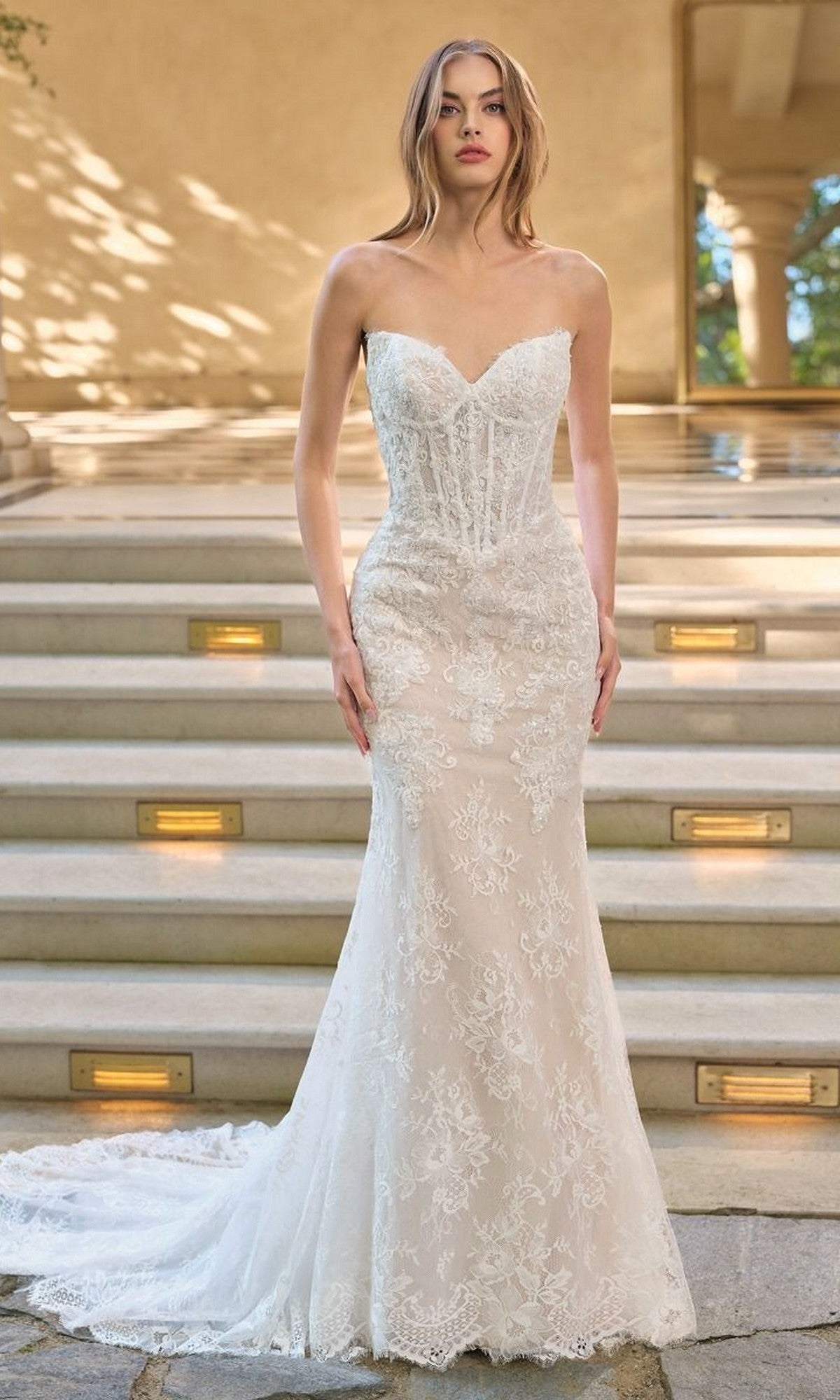 A woman stands on stone steps wearing the Andrea and Leo WL071 long wedding dress, featuring a strapless sweetheart neckline, white lace fabric, and a long train.