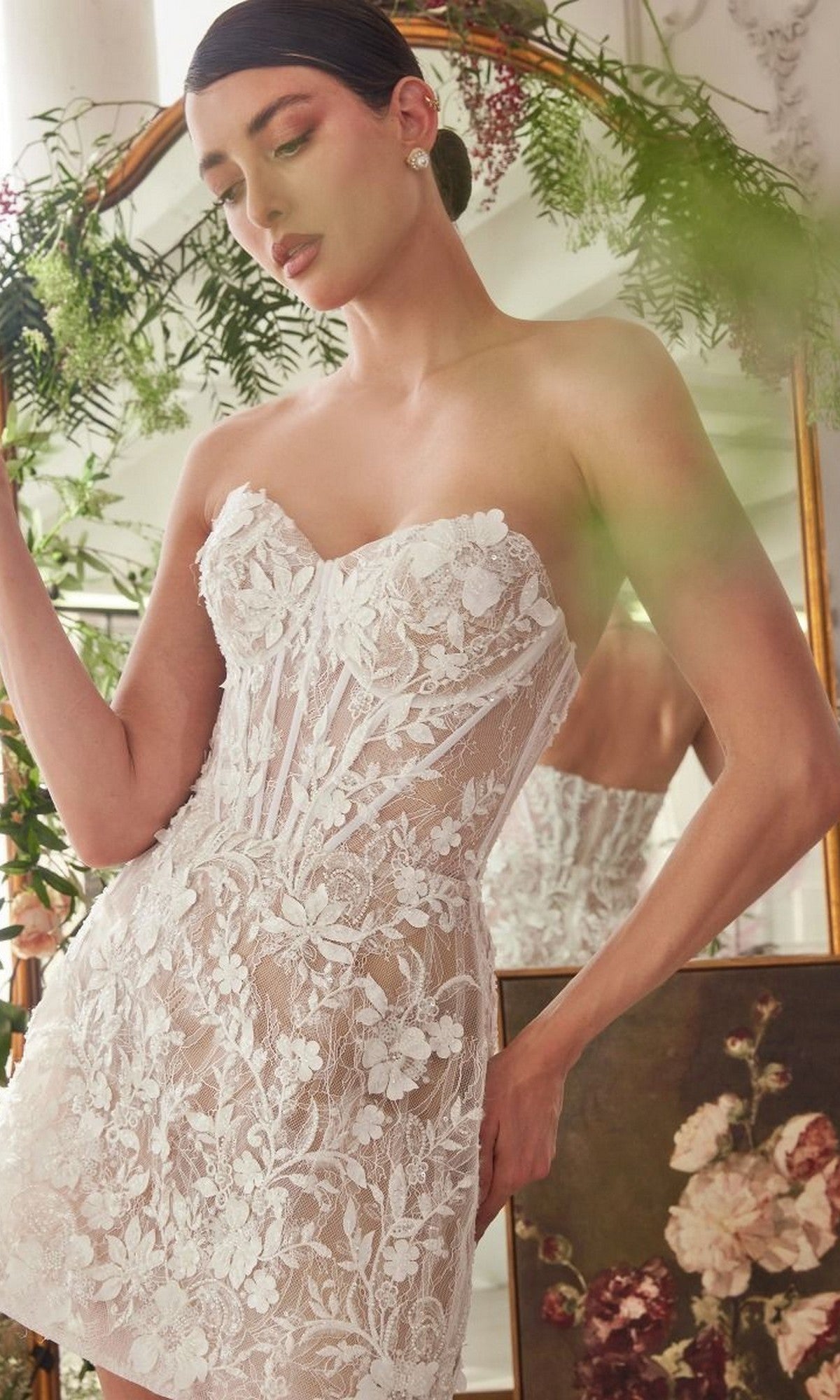 A woman stands in front of a mirror with flowers and greenery, wearing the Andrea and Leo WL084 Short White Dress featuring a strapless design, sheer white floral embroidery, and delicate floral appliques.