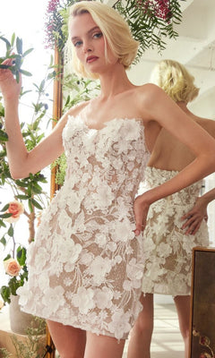 A woman wearing the Andrea and Leo WL085 Short White Dress, a strapless design with 3D floral lace appliqués and a sheer silhouette, stands indoors near a mirror and plants.