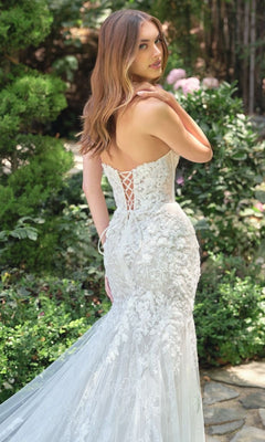 A woman in the Andrea & Leo WL088 strapless mermaid bridal dress with a lace-up back stands outdoors on a stone path, surrounded by greenery.