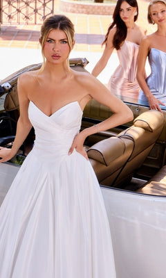Woman in the A-Line Long Classic White Dress: Ladivine WN015, featuring a strapless sweetheart neckline, stands by a convertible car as two women in pastel dresses appear in the background.