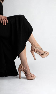 A person in a black dress sits cross-legged, highlighting the Wren Nude Metallic Prom Shoes by Touch Ups 4503, featuring a strappy design and high heels against a plain background.