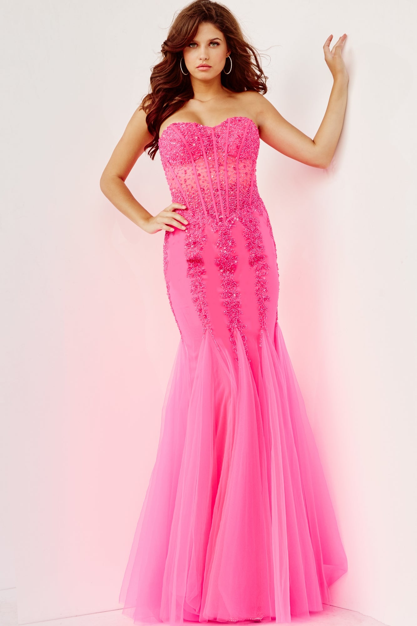 A woman wears the Long Strapless Sweetheart Jovani Prom Dress 5908, a floor-length, bright pink mermaid gown with sequin embellishments, standing against a white wall.