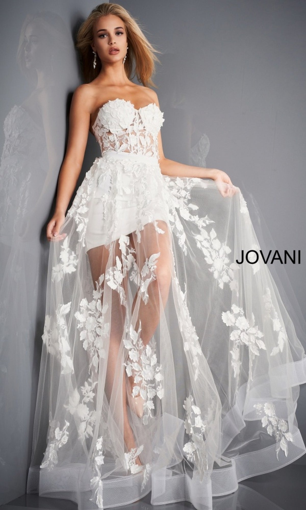 A woman models the Jovani Strapless Sweetheart Sheer Corset Prom Dress, a strapless white gown with a sheer floral overlay and short lining, against a gray background. 
