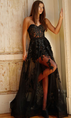 A woman stands indoors wearing the Jovani Strapless Sweetheart Sheer Corset Prom Dress, a floor-length black gown with floral details, posing with one hand on a doorframe.