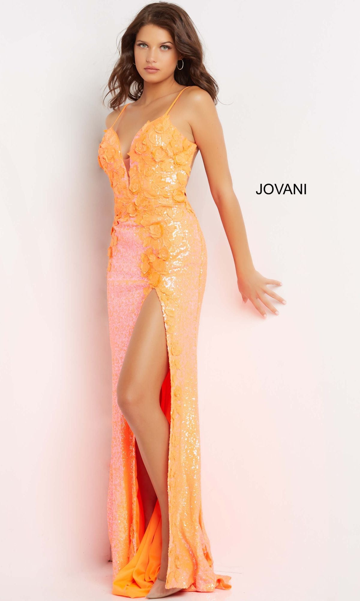 A woman in a fitted, orange Long Sequin Jovani Prom Dress with appliques and a high slit stands against a white background. The word 