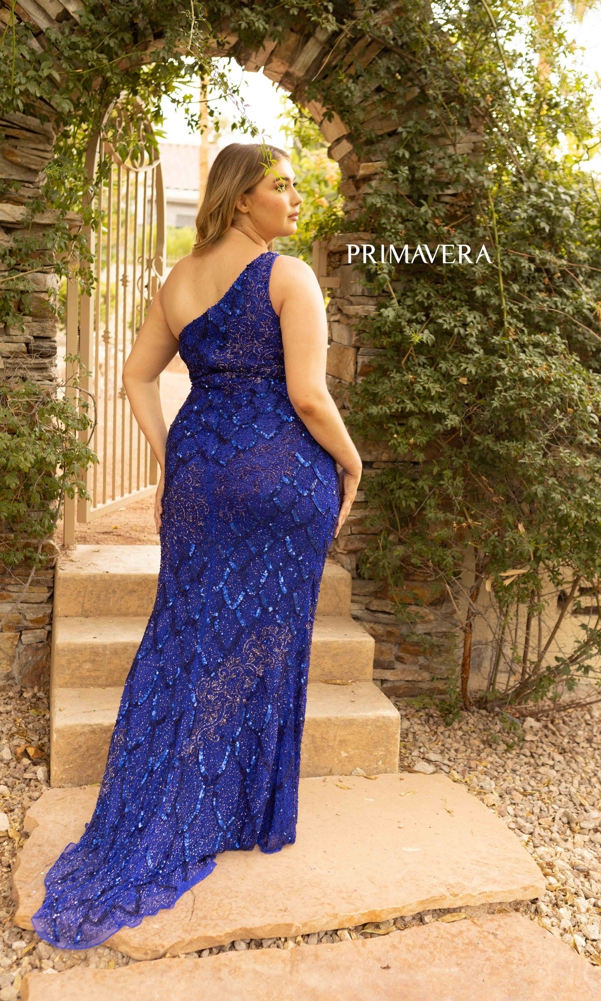 A woman in the One-Shoulder Sequin Plus-Size Prom Dress 14002 stands on stone steps beneath a vine-covered archway, her glittering blue gown catching the light as 