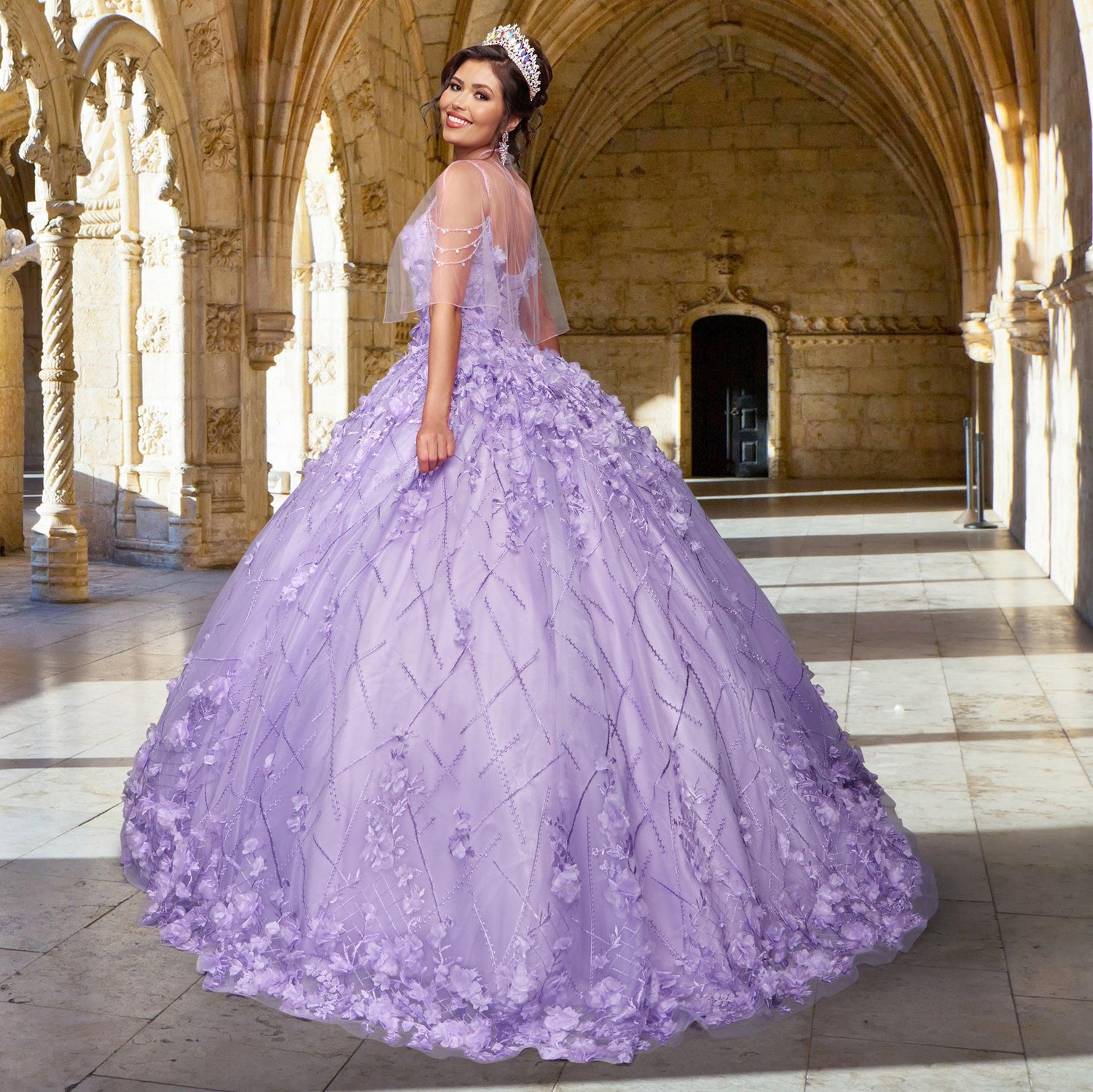 A woman in the Quinceañera Sample Dress QF105 and a tiara stands in an arched stone hallway, looking over her shoulder with a smile.