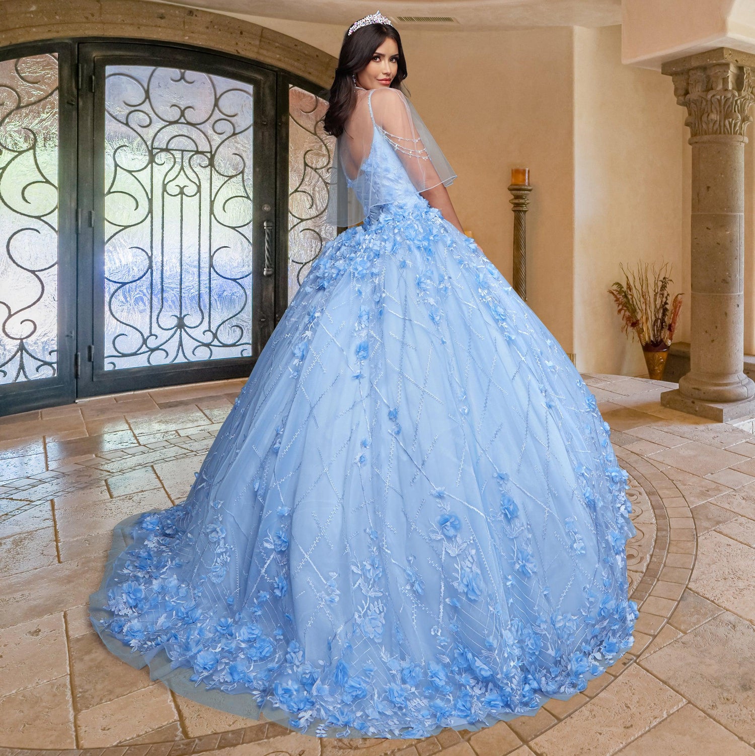 A woman models the Quinceañera Sample Dress QF105, a light blue, embellished gown, standing indoors on a tiled floor and glancing over her shoulder—an ideal choice for your special celebration.