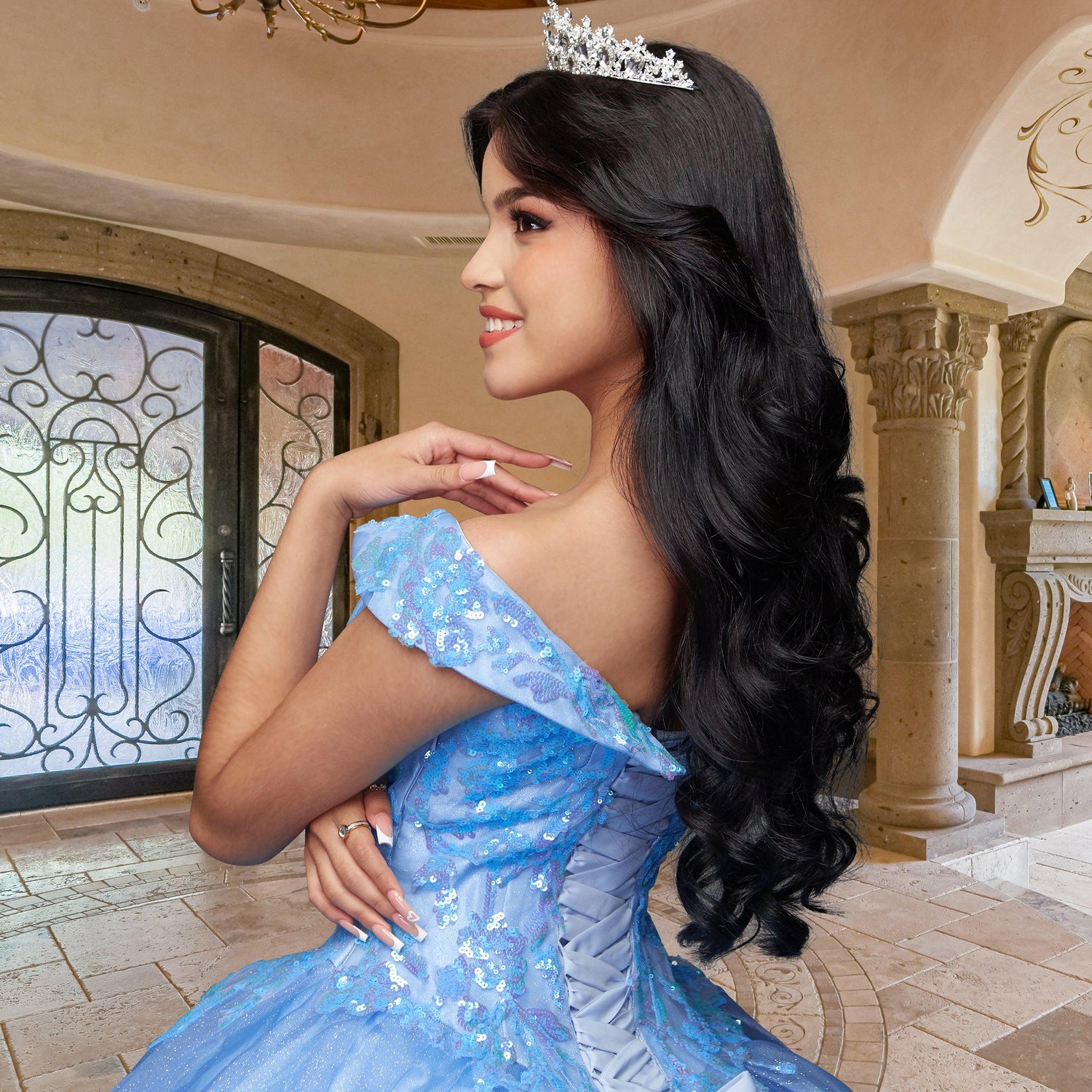 A young woman poses indoors, smiling in the ornate, well-lit room while wearing the Quinceañera Sample Dress QF104—a blue, off-the-shoulder gown—paired with a tiara and her hand resting on her shoulder.