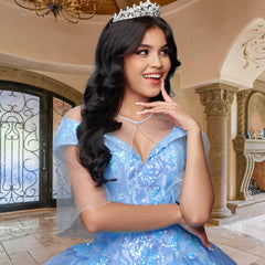 A young woman wears the Quinceañera Sample Dress QF104, a blue, sparkly off-shoulder gown with a tiara, posing indoors with a smile in an elegant room featuring arched doorways and ornate decor.
