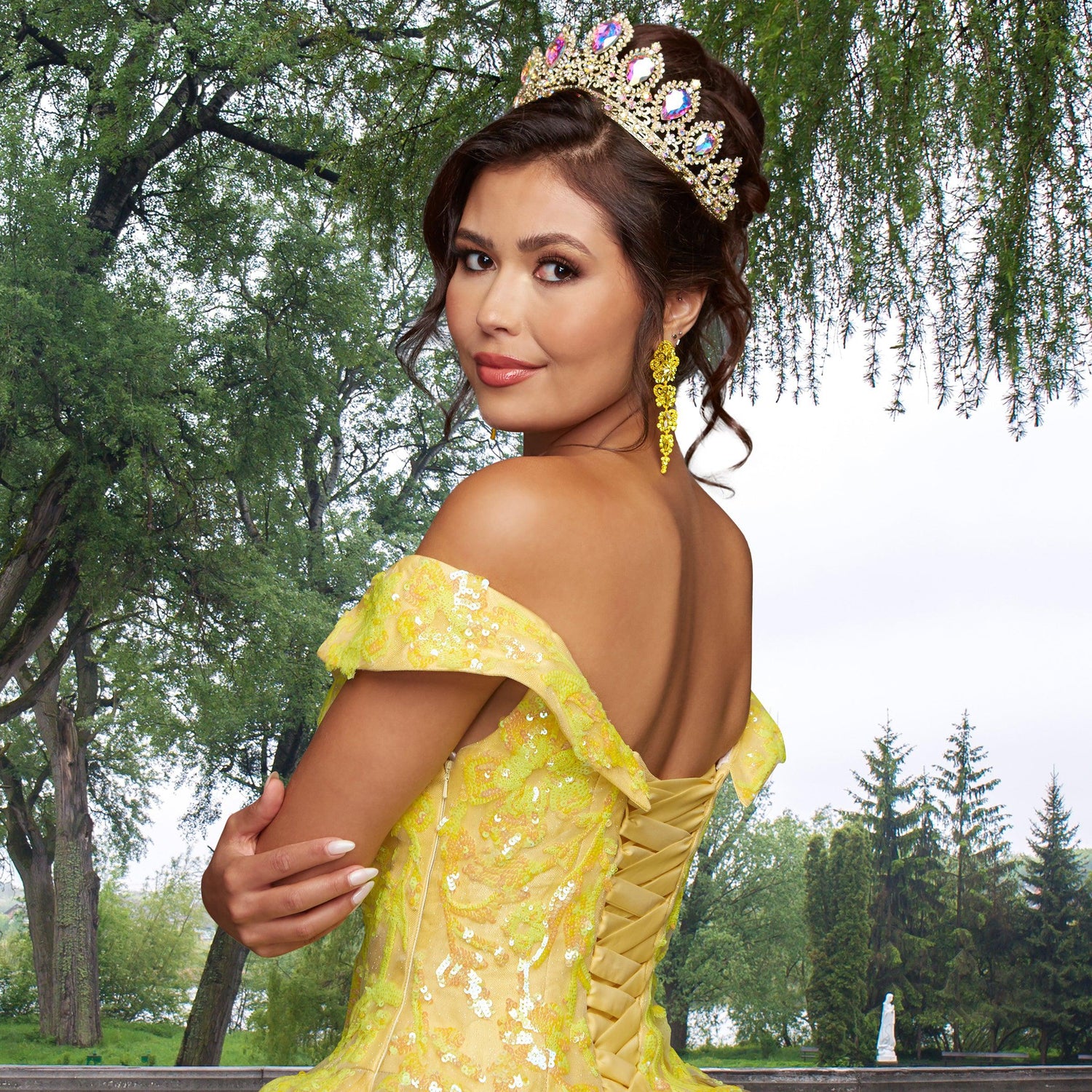 Woman wearing the Quinceañera Sample Dress QF104 and a jeweled tiara poses outdoors, surrounded by lush greenery and trees.