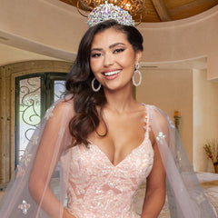A woman indoors smiles in the Quinceañera Sample Dress QF106, a beaded light pink ball gown. She wears a jeweled crown, large earrings, and has long, dark wavy hair draped over one shoulder.