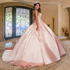 A young woman wearing the Quinceañera Sample Dress QF106 and a tiara stands in a decorated room with arched doors and stone columns, smiling at the camera.