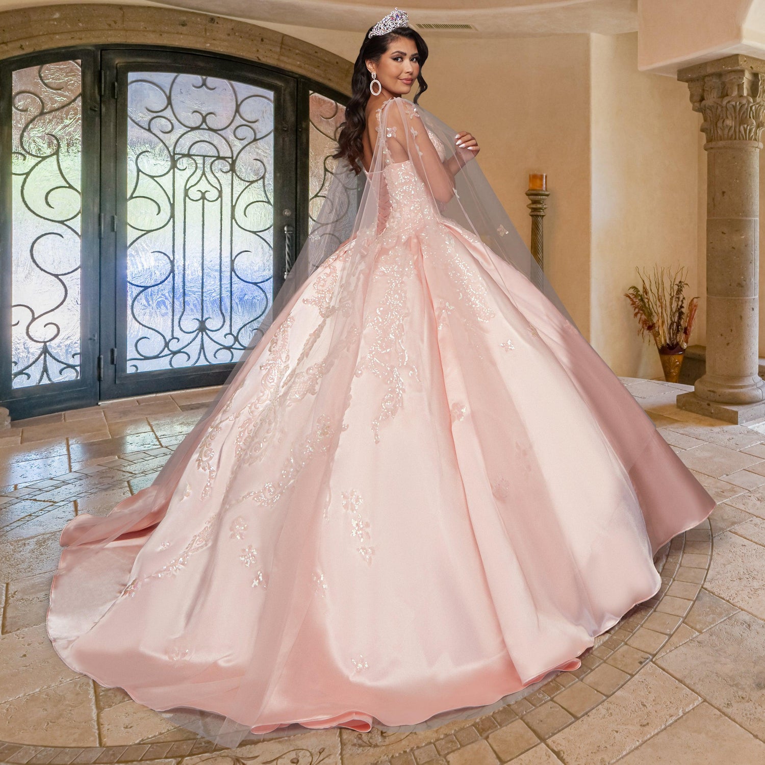 A woman wearing the Quinceañera Sample Dress QF106, a pink sequin gown and tiara, stands in an elegant room with stone tile flooring and decorative columns, facing away from the camera.