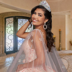 A woman wearing the Quinceañera Sample Dress QF106—featuring a sparkling tiara and pink ball gown with sheer, embroidered sleeves—smiles indoors at the camera.
