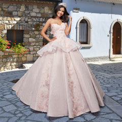 A woman in the Quinceañera Sample Dress QF115 and a tiara smiles on a cobblestone street, standing before a stone and stucco building.