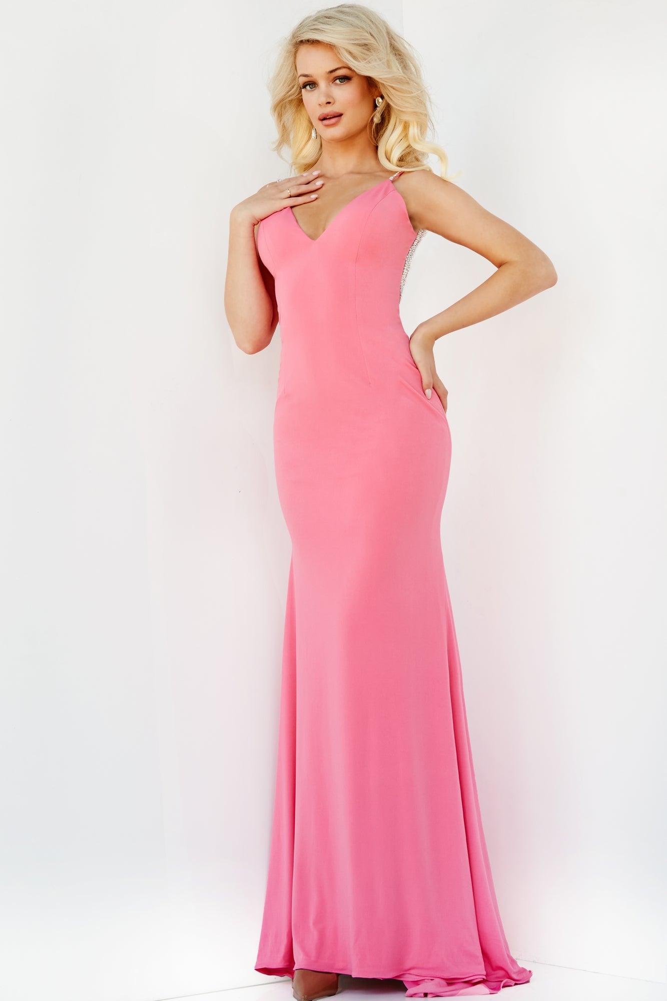 A woman stands against a white background wearing the Fitted Backless Long Jovani Prom Gown 07297: a long, fitted, sleeveless pink dress with a V-neckline and side slit.