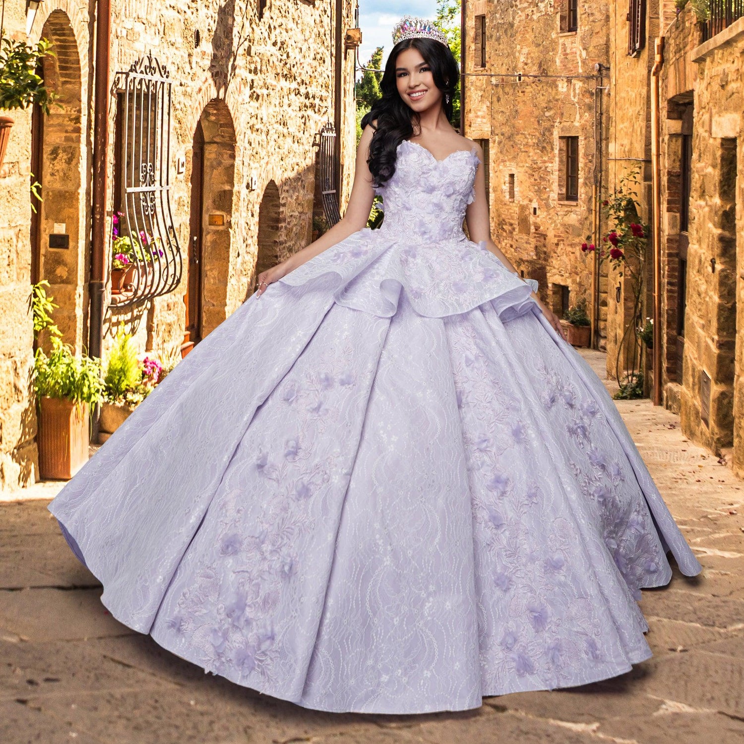A young woman wearing the Quinceañera Sample Dress QF115—a light purple, strapless gown with a tiara—stands smiling on a cobblestone street lined with old stone buildings.