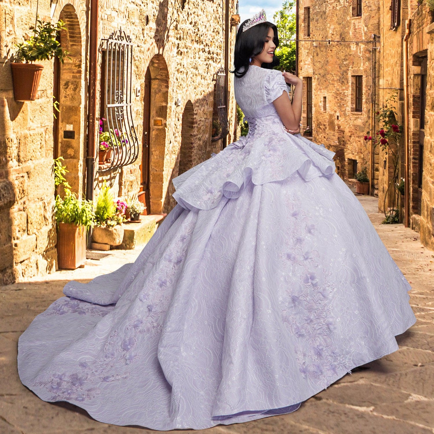A woman in a Quinceañera Sample Dress QF115 and tiara stands smiling in a sunlit, narrow stone street lined with rustic buildings and potted plants.