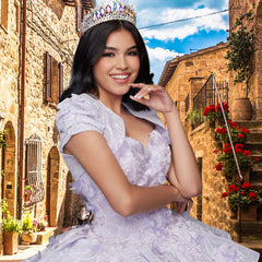 A woman in the Quinceañera Sample Dress QF115 and a jeweled crown smiles while posing on a rustic stone street adorned with flowers beneath a blue sky.