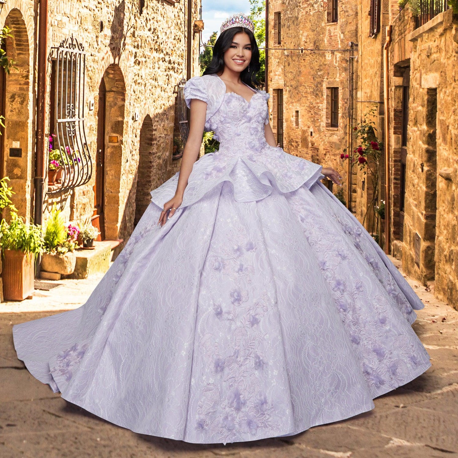 A young woman in the Quinceañera Sample Dress QF115, a light purple ornate sweetheart gown with a tiara, smiles while standing on a narrow sunlit stone street adorned with flowers and plants.