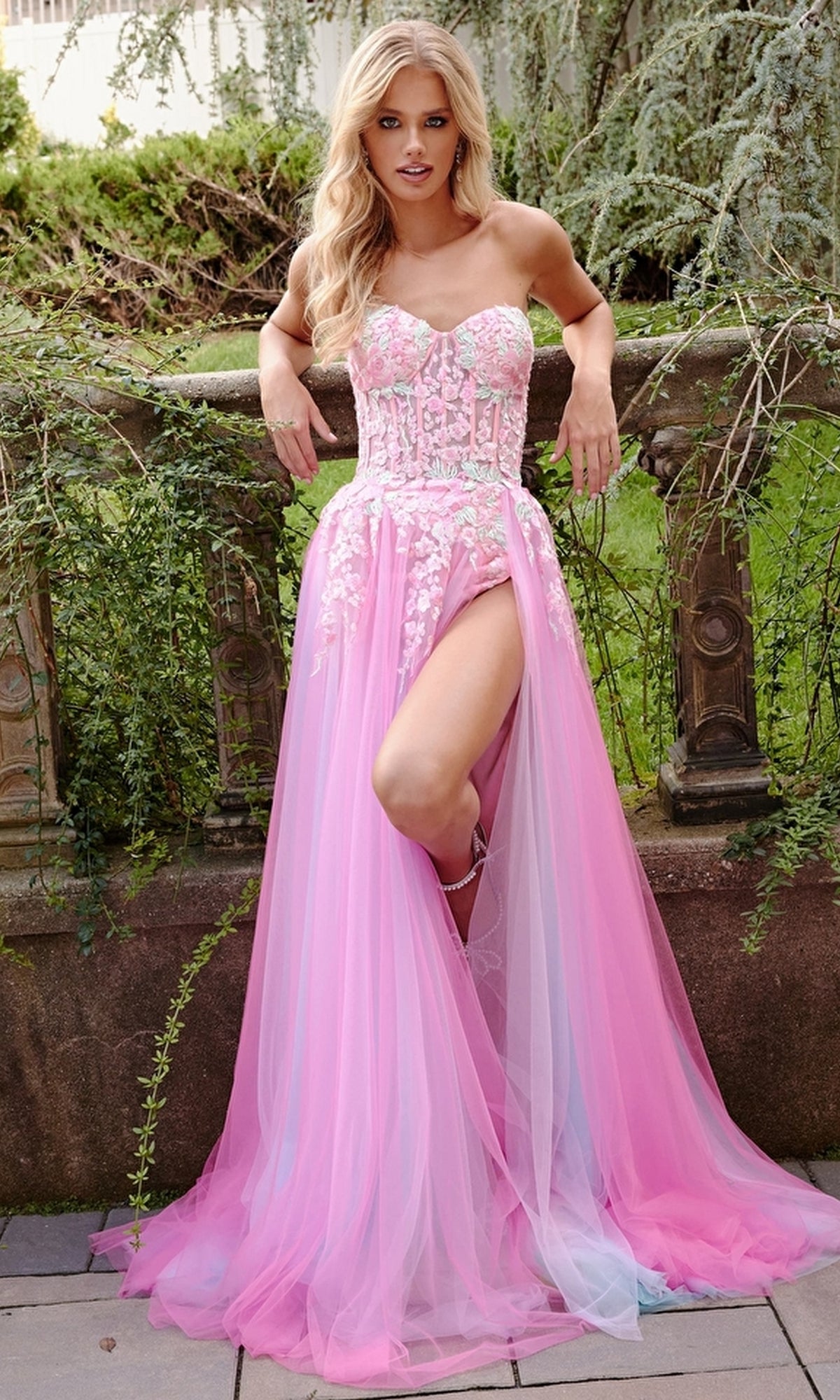 A woman wearing the Jovani Sheer-Corset Prom Dress with floral embroidery and a pink-lavender sheer skirt with a high slit stands outdoors against a stone railing.