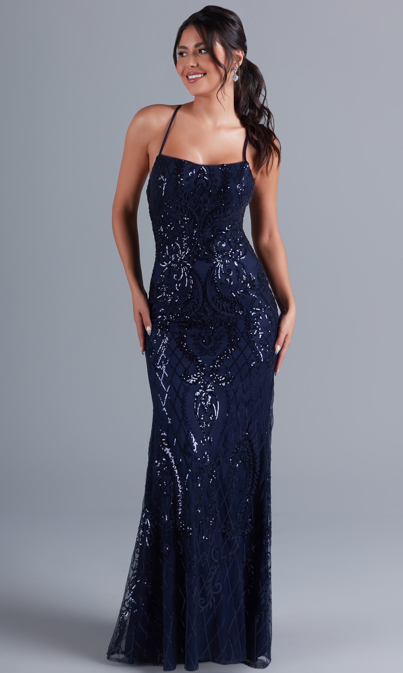 A woman stands against a plain gray background, smiling with her hands at her sides, wearing the PromGirl Statement-Back Long Sequin Prom Dress in navy blue with thin straps and a floor-length silhouette.