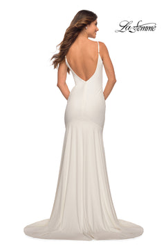 A woman in a white, floor-length La Femme Backless Long Prom Dress with a train, deep V-back, and thin straps stands with her back to the camera. The La Femme logo appears in the upper right corner.
