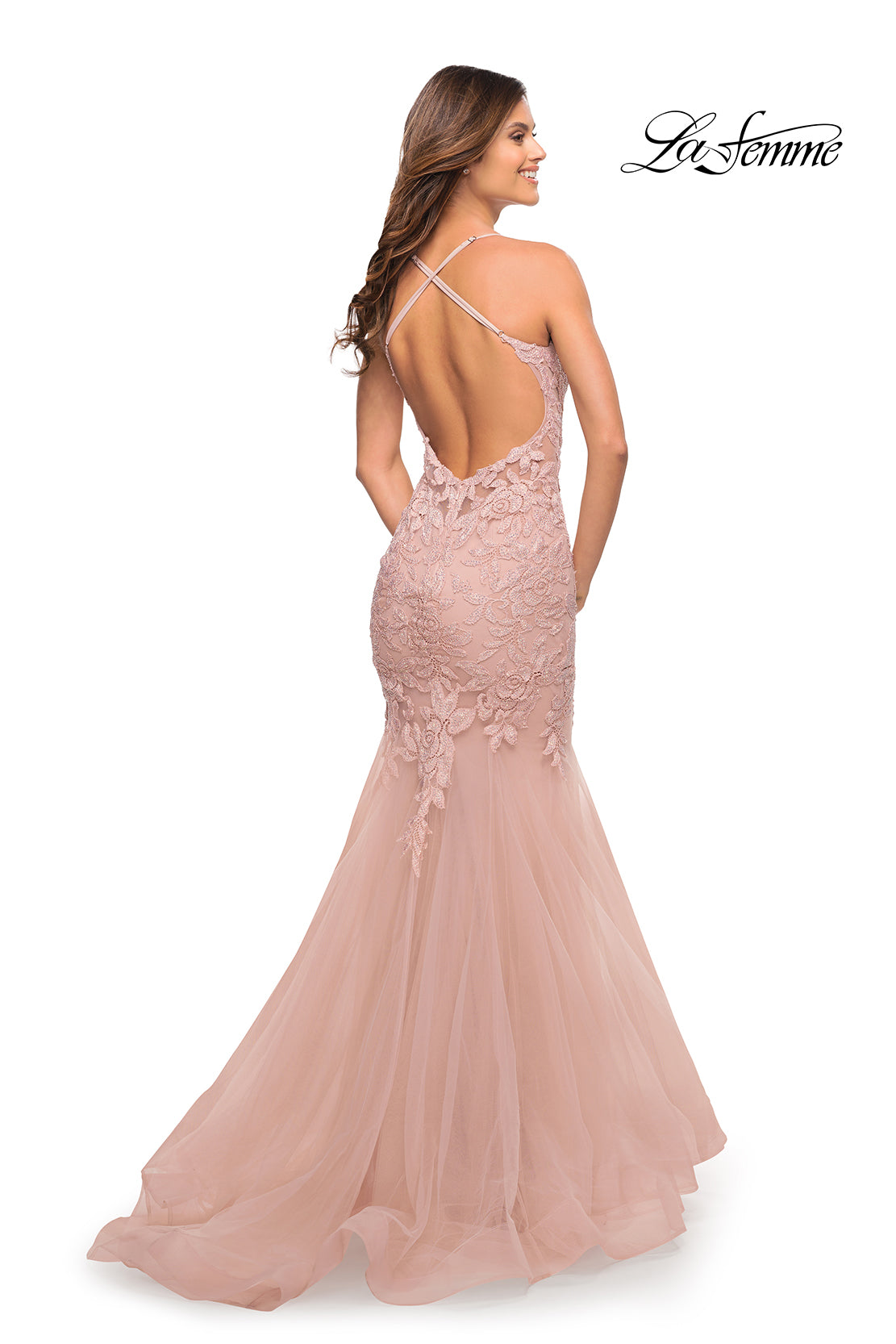 A woman stands facing away, wearing the Backless Long Lace Mermaid Prom Dress by La Femme in blush pink. The formal gown features delicate lace, crisscross straps, a mermaid silhouette, and the 