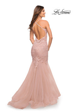 A woman stands facing away, wearing the Backless Long Lace Mermaid Prom Dress by La Femme in blush pink. The formal gown features delicate lace, crisscross straps, a mermaid silhouette, and the "La Femme" logo in the upper right corner.