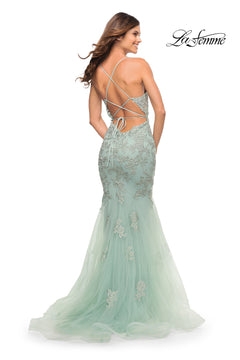 A woman models the La Femme Long Lace Designer Mermaid Prom Dress in light green, featuring a fitted bodice, open back, and crisscross straps. She stands looking over her shoulder; "La Femme" logo appears in the top right.